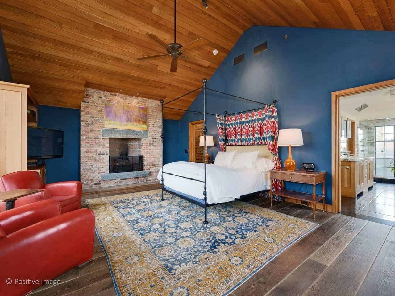 This industrial-style primary bedroom has the warm embrace of a traditional fireplace that is inlaid with red bricks. This stands out against the blue walls but complements the wooden cathedral ceiling that has a ceiling fan over the four-poster bed.